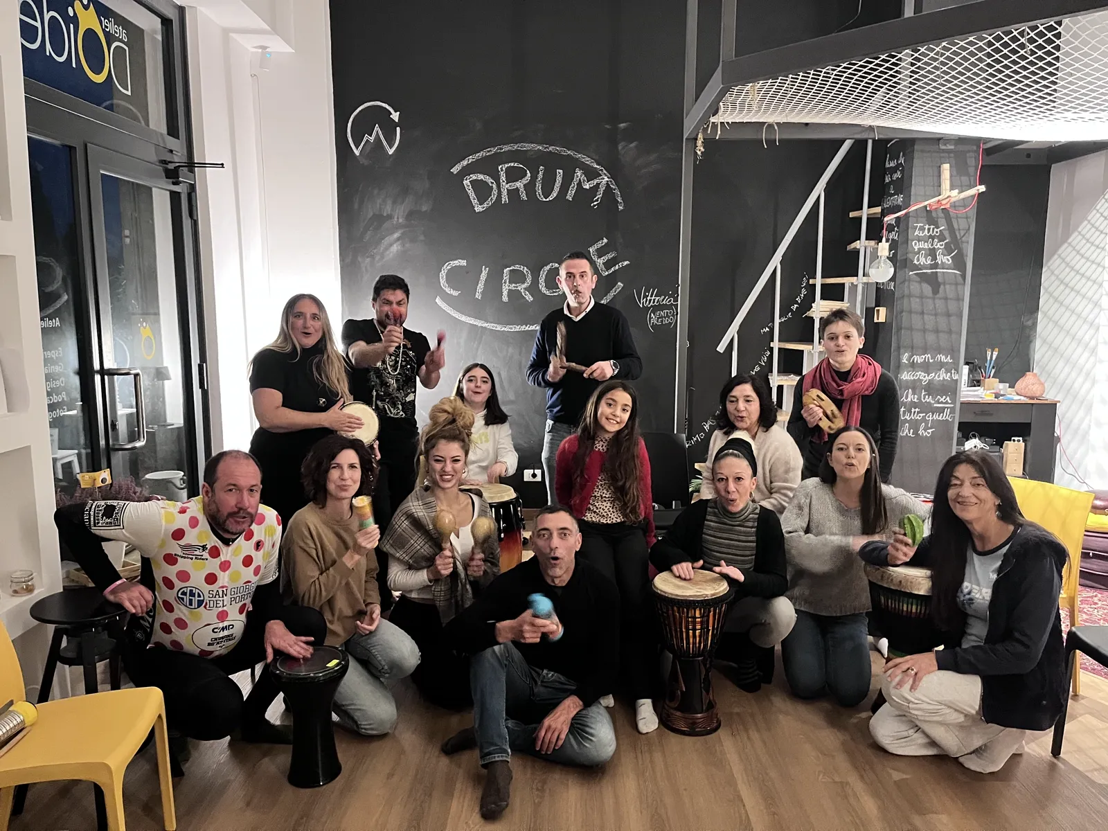 Una quindicina di persone in cerchio con tamburi e percussioni, sorridenti, durante un drum circle in atelier