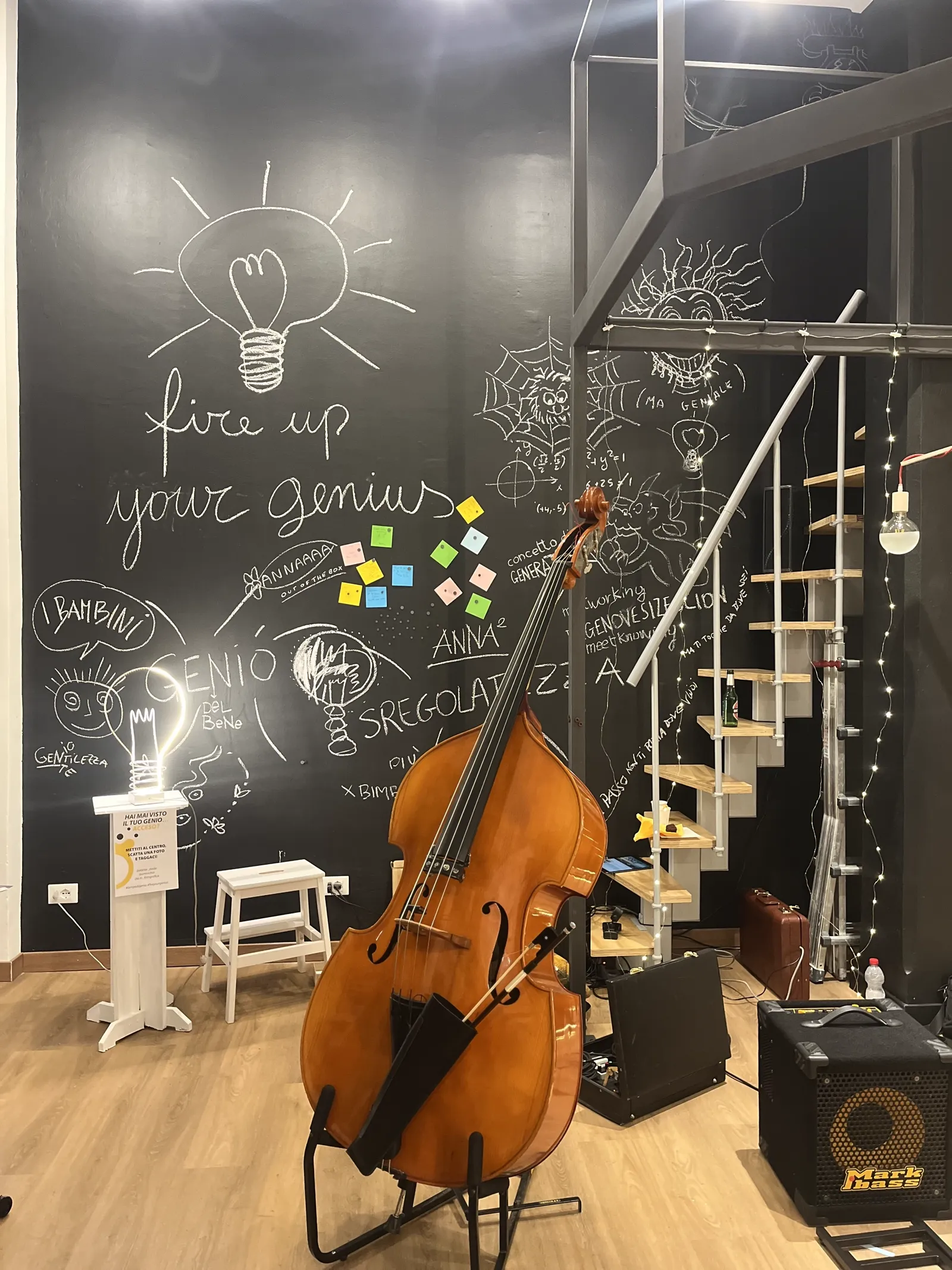 Parete lavagna nera con la scritta a gesso 'fire up your genius', un contrabbasso in primo piano e una scala a chiocciola