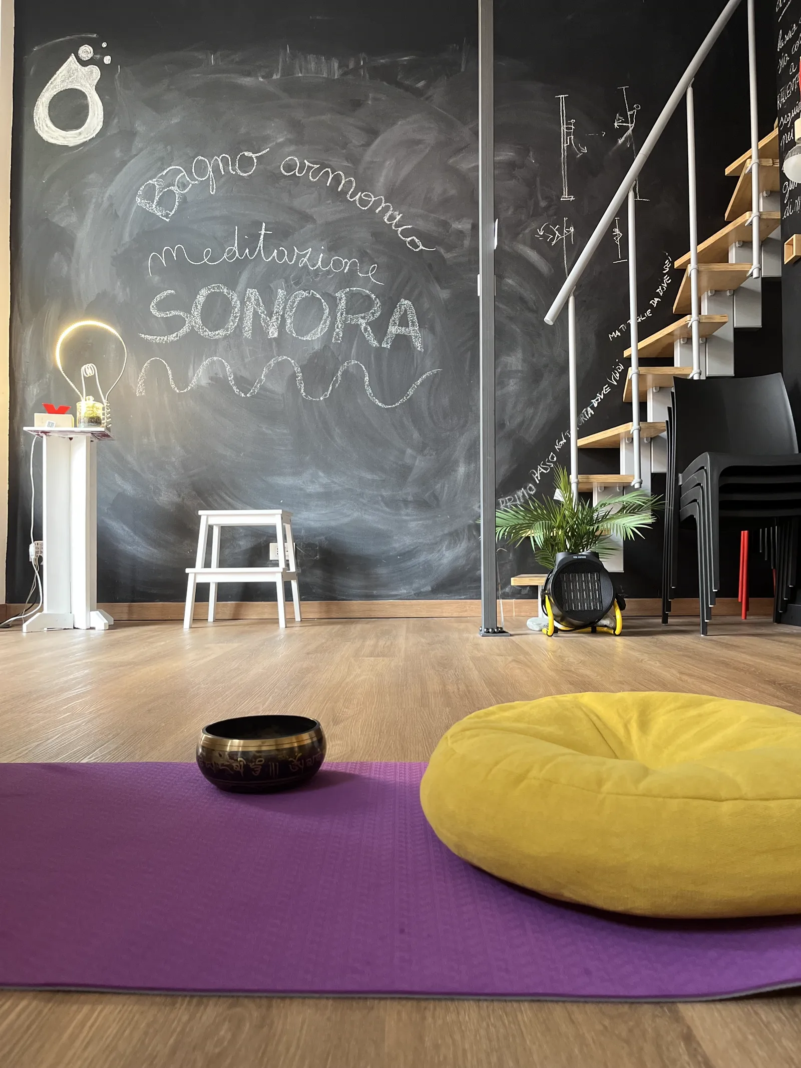Tappeto viola con campana tibetana e pouf giallo, parete con scritta a gesso 'bagno armonico, meditazione sonora'