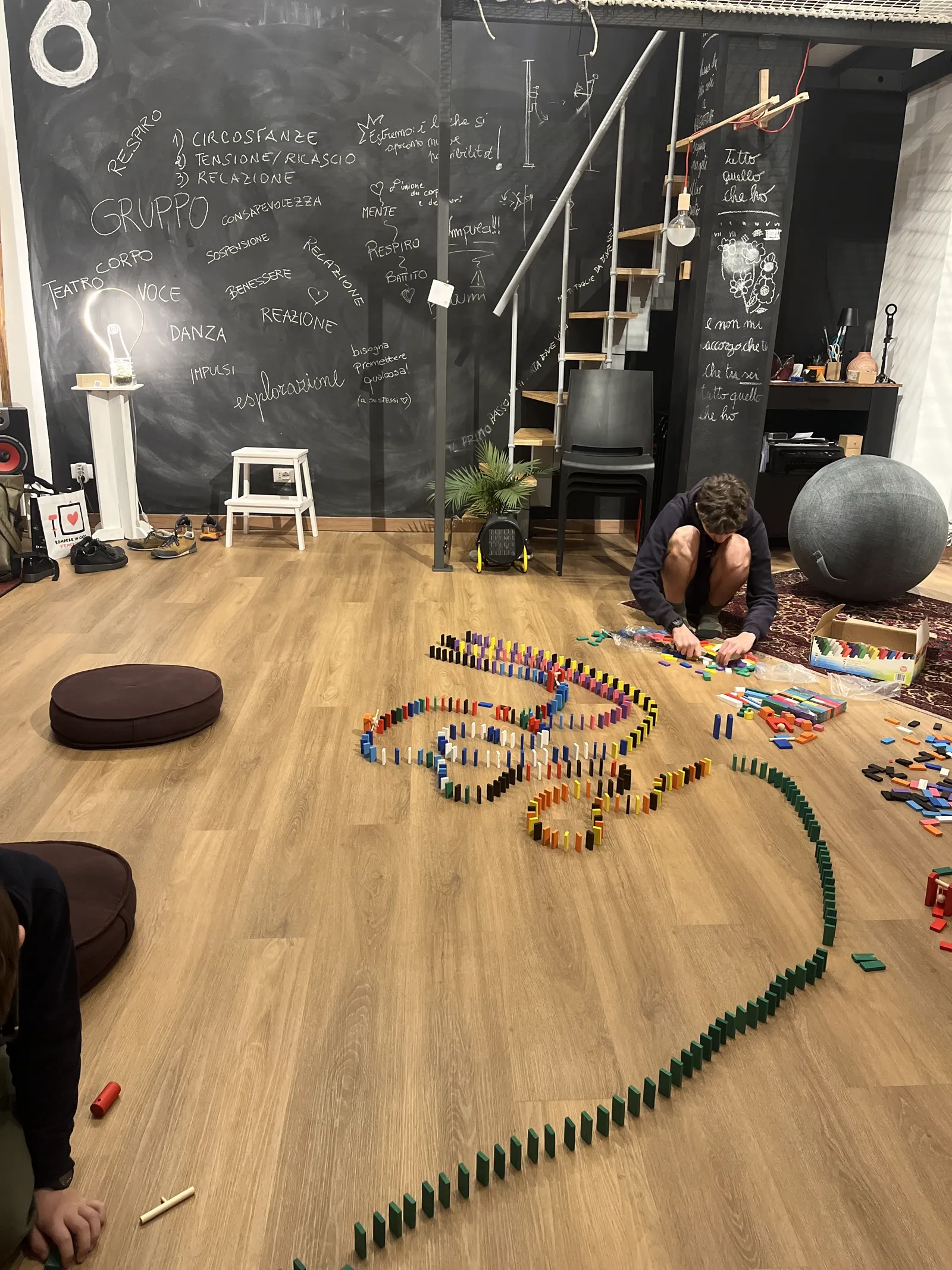 Una persona costruisce una grande forma con tessere di domino colorate sul pavimento, davanti a una parete lavagna con la scritta GRUPPO e parole creative