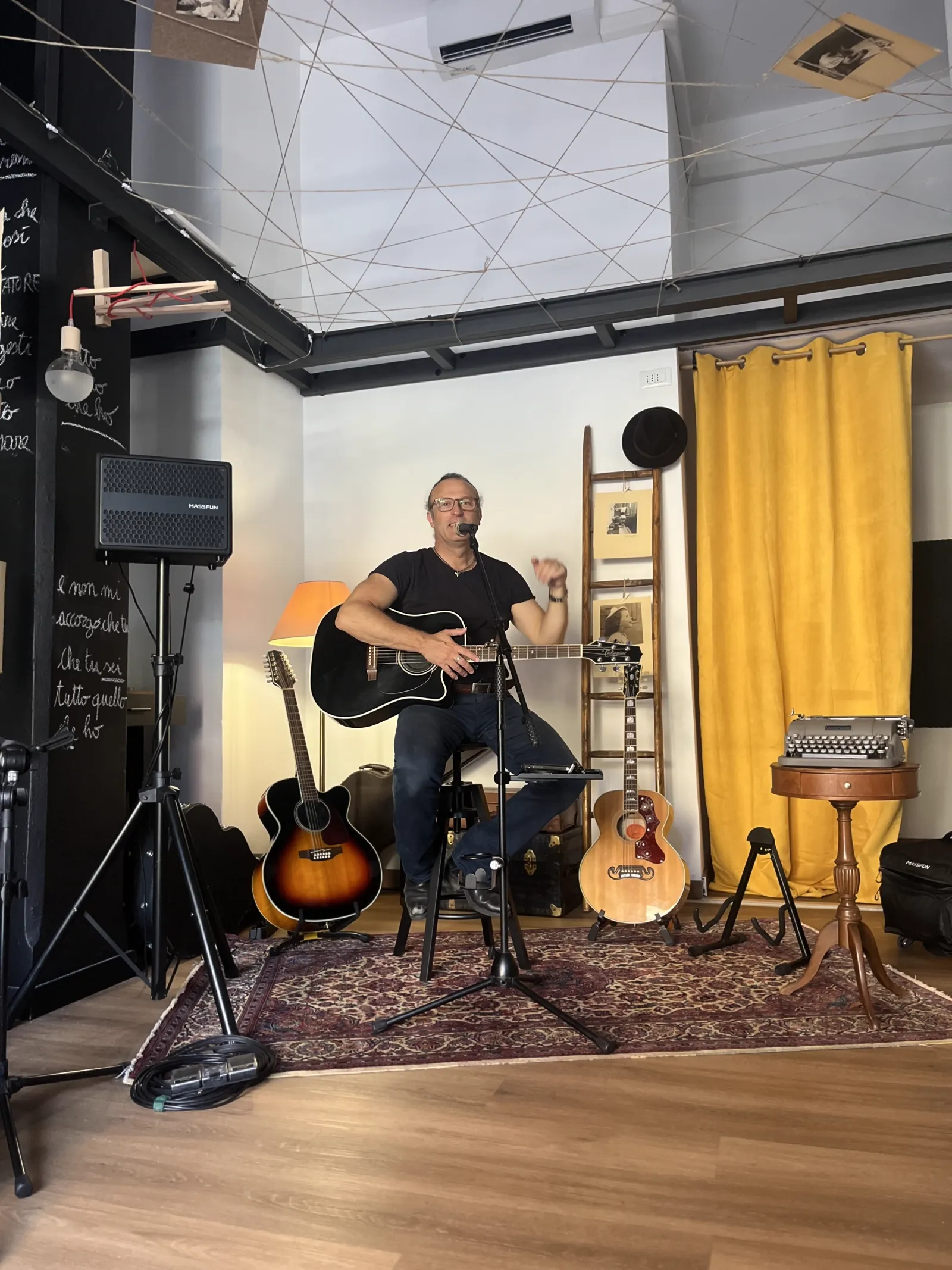Un musicista con la chitarra acustica in atelier, davanti a una grande tenda gialla, tappeto persiano, lampada vintage
